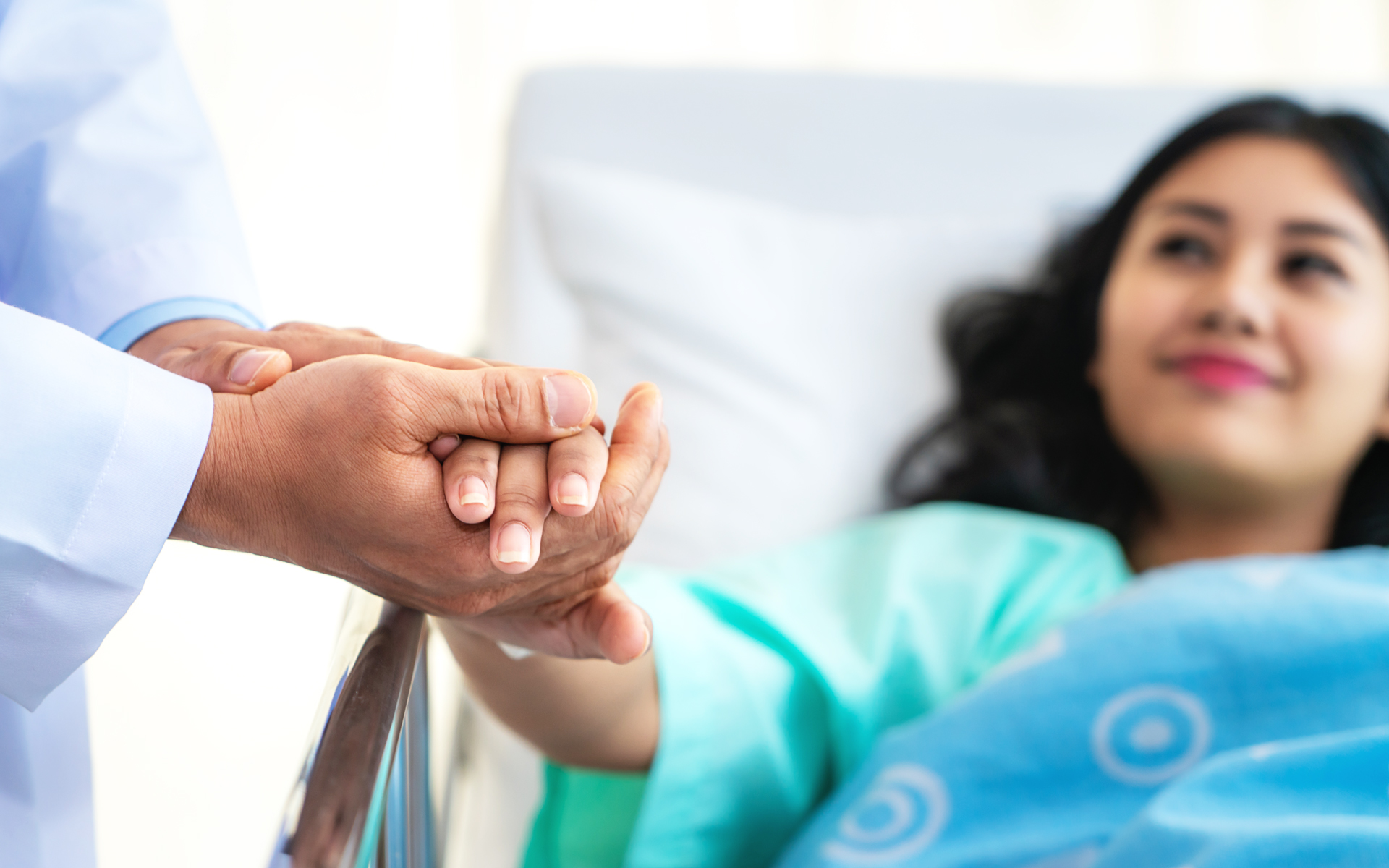 Image for Best Tips on How to Avoid or Manage Medical Debt. Image of a doctor holding a patient's hand with both of their hands while the patient lies in bed.