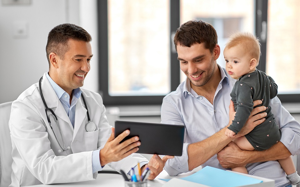 Image for 11 The biggest health stories to watch in 2023. A doctor shows a man with a baby something on a tablet.